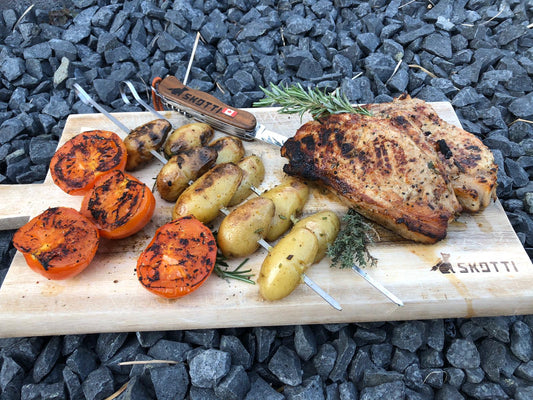 Kalbskotelett vom Grill angerichtet auf einem Holzbrett mit Grilltomaten und Rosmarin-Kartoffelspießen