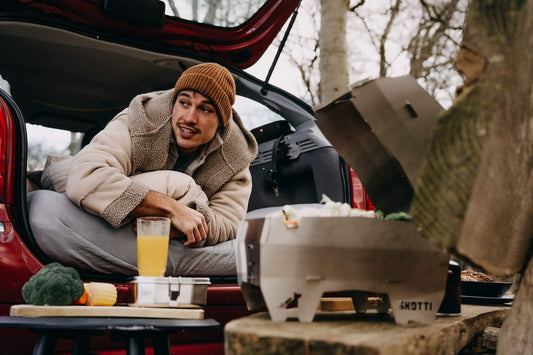 Junger Outdoor-Lover liegt auf einer Matraze in seinem Auto und grillt in der Natur auf dem SKOTTI Grill